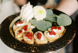 A close-up view of a cater waiter holding a silver platter with eucalyptus and white flowers as decor, and cream cheese and roasted red pepper crostinis