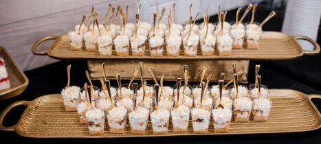 Two long bronze trays with numerous individual dessert shooters of salted caramel cheesecake parfaits